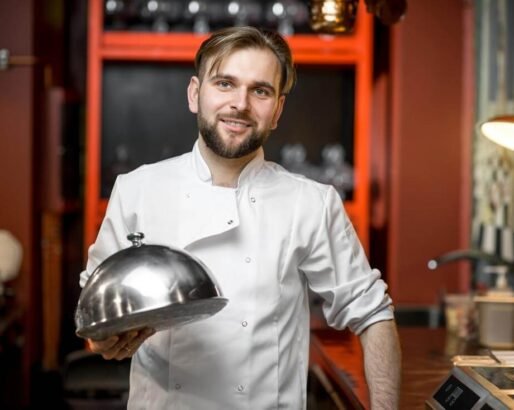 Chef cook portrait in uniform with steel cloche at the red restaurant kitchen interior. Image with small deph of field and blurred background